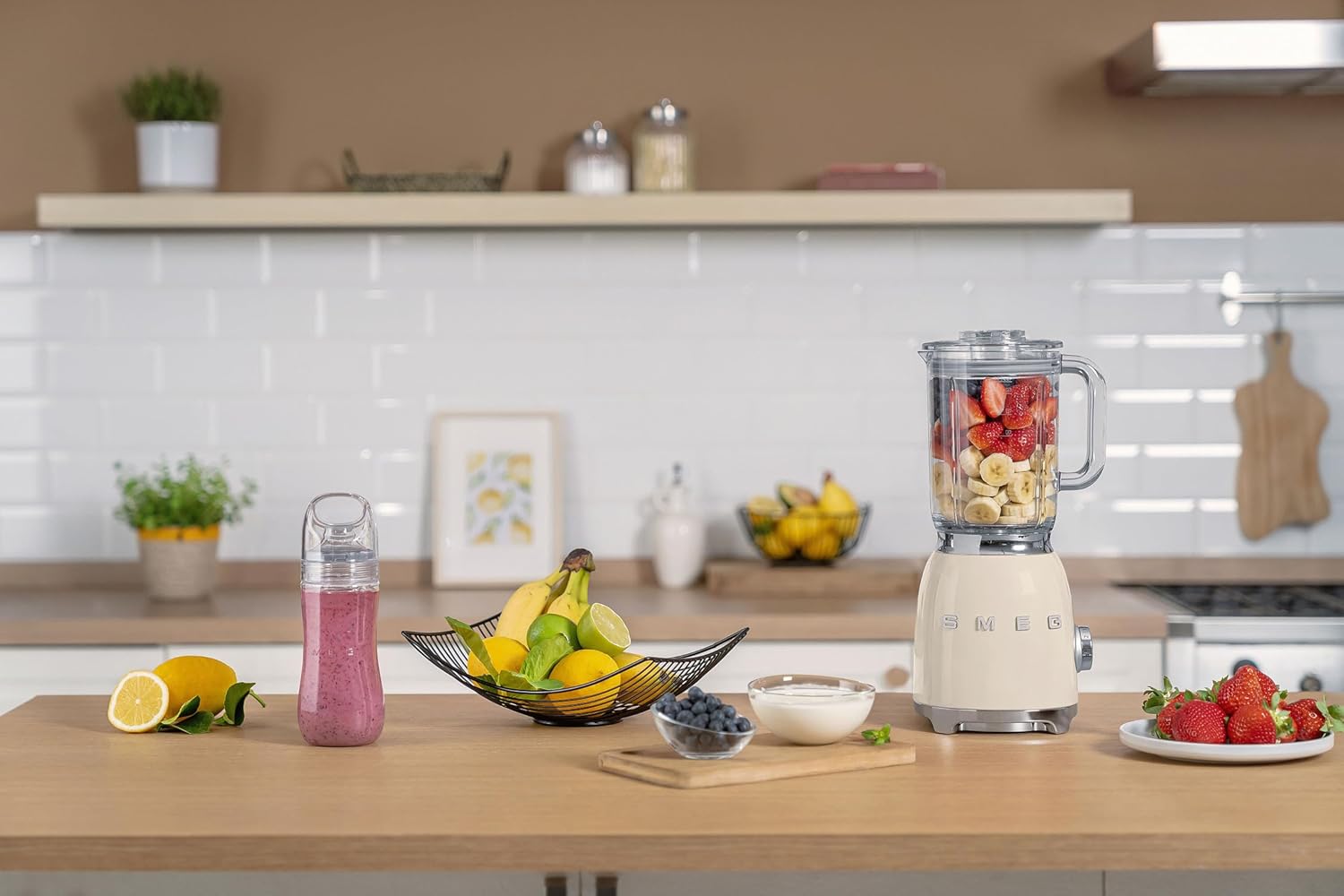 SMEG BLF03 blender i kremfarge på en kjøkkenbenk, fylt med jordbær og bananer, omgitt av frukt, yoghurt, og en ferdiglaget smoothie i bærbar flaske. Smeg blender