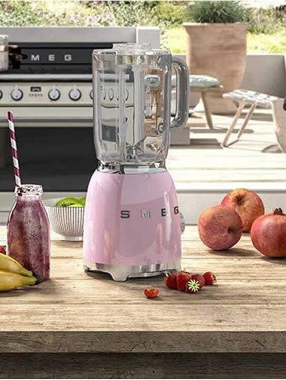 Smeg BLF01PKEU blender i pastellrosa på en kjøkkenbenk, omgitt av fersk frukt og en smoothieflaske med sugerør, ideell for sunn matlaging.