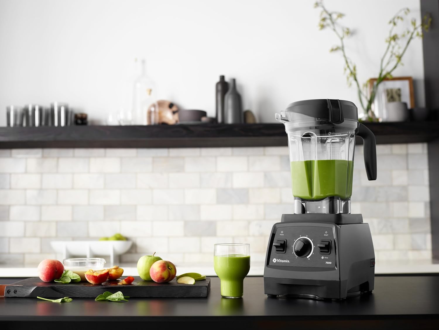 Vitamix 7500 blender på et kjøkken med en grønn smoothie i beholderen og fersk frukt på skjærebrett ved siden av.